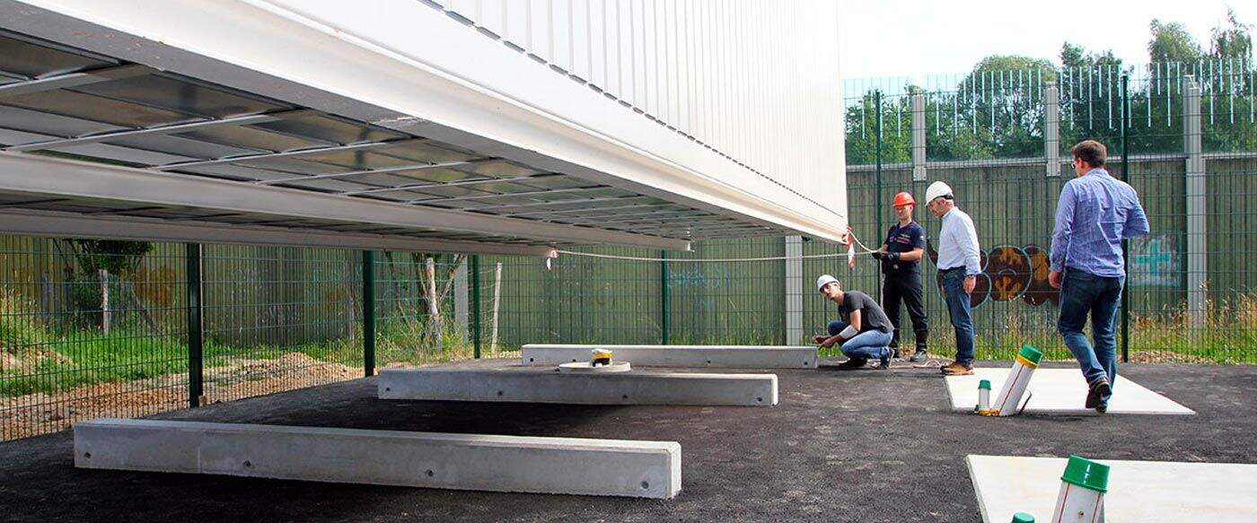 Travailleurs sur un chantier, un bâtiment suspendu en métal au-dessus de fondations en béton, entouré de grillages et d'arbres.