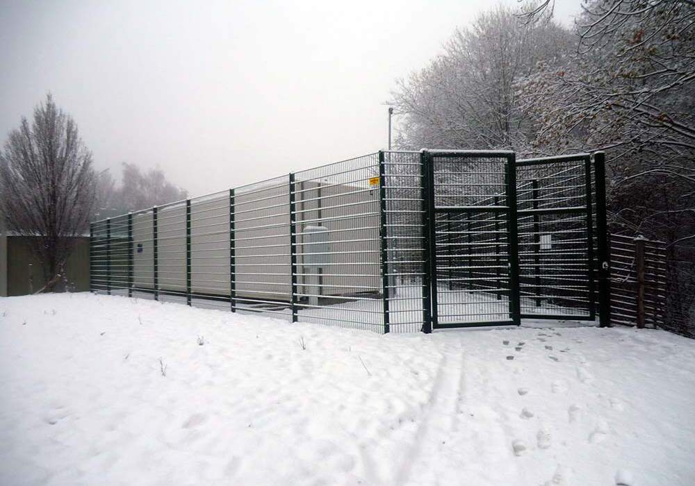 Clôture en métal vert entourant un espace, recouvert de neige, avec des arbres en arrière-plan sous un ciel brumeux.