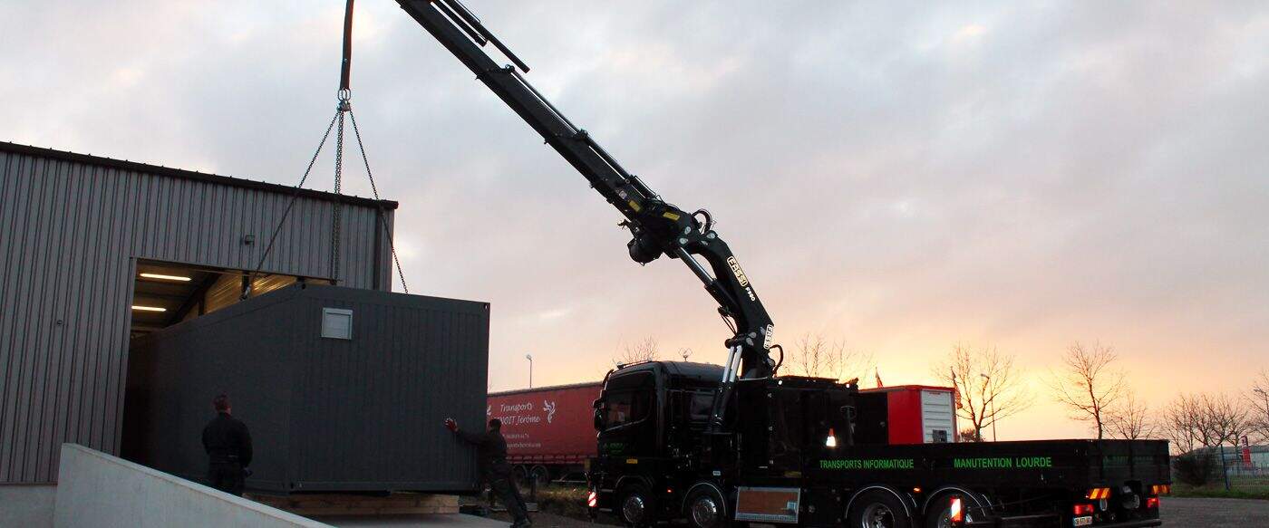 Grue noire en action soulevant un conteneur gris devant un entrepôt, sous un ciel légèrement nuageux à l'aube.