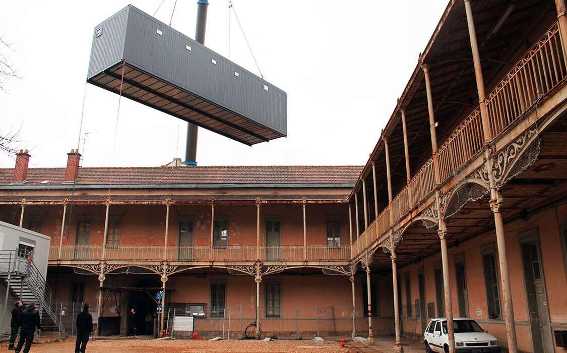 Conteneur grise suspendu au-dessus d'une cour intérieure avec des structures métalliques ornées, un bâtiment ancien en arrière-plan.