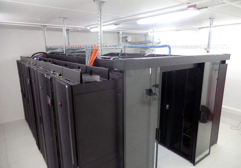 Salle de serveur avec des racks noirs, câbles colorés suspendus, sol en carrelage clair et éclairage fluorescent.