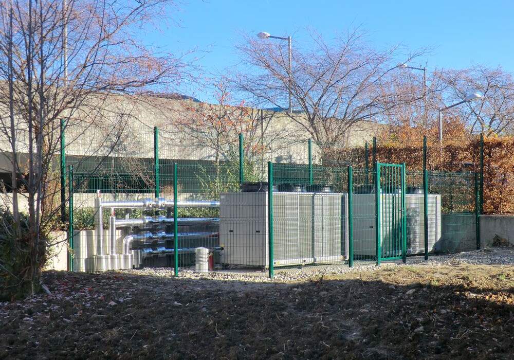 Installation de climatisation servere, entourée de clôtures vertes, avec des arbres à feuilles caduques et un ciel bleu en arrière-plan.