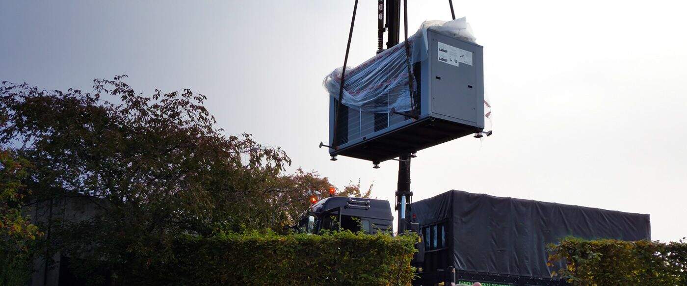 Un container gris est suspendu par une grue, entouré d'arbres et d'un camion à l'arrière-plan sous un ciel nuageux.