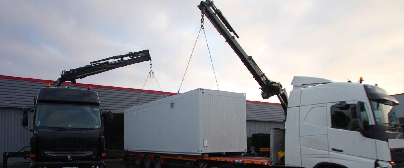 Camion lourd avec une grue soulevant un conteneur blanc sur une remorque, devant un bâtiment aux murs gris et un ciel nuageux.