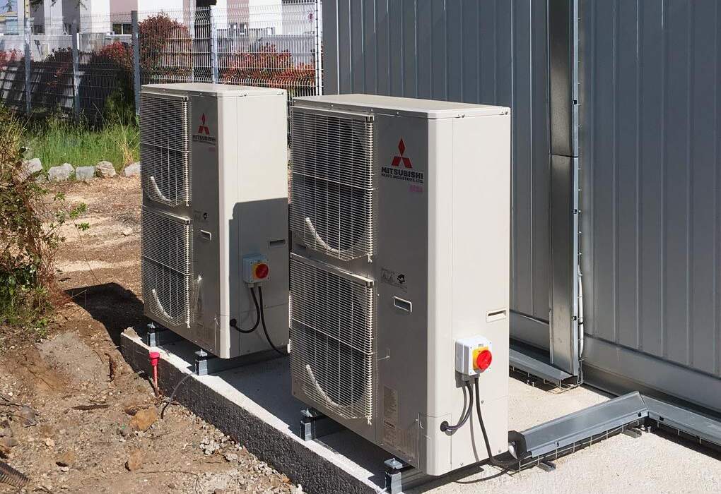 Deux climatiseurs Mitsubishi beige installés à l'extérieur, sur une base en béton, avec un arrière-plan de clôture et d'arbres.