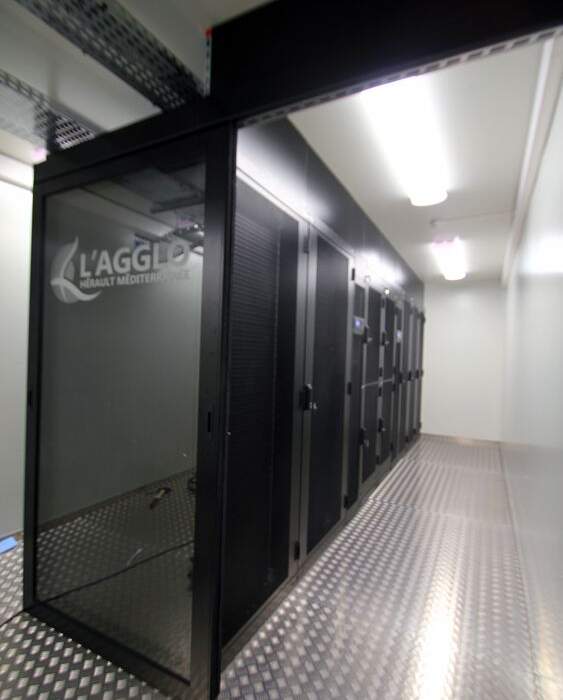 Salle serveur moderne avec des racks de matériel informatique, murs noirs et sol en métal texturé, éclairage fluorescent au plafond.