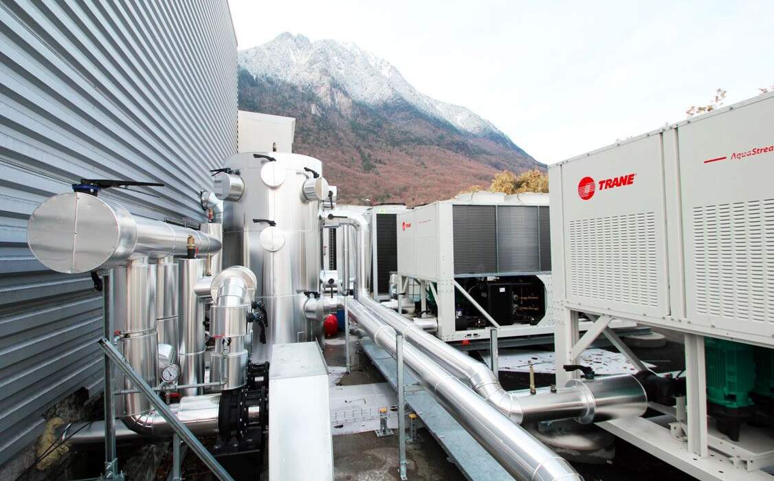 Système de réfrigération avec conduits métalliques, unités Trane et montagnes enneigées en arrière-plan sous un ciel clair.