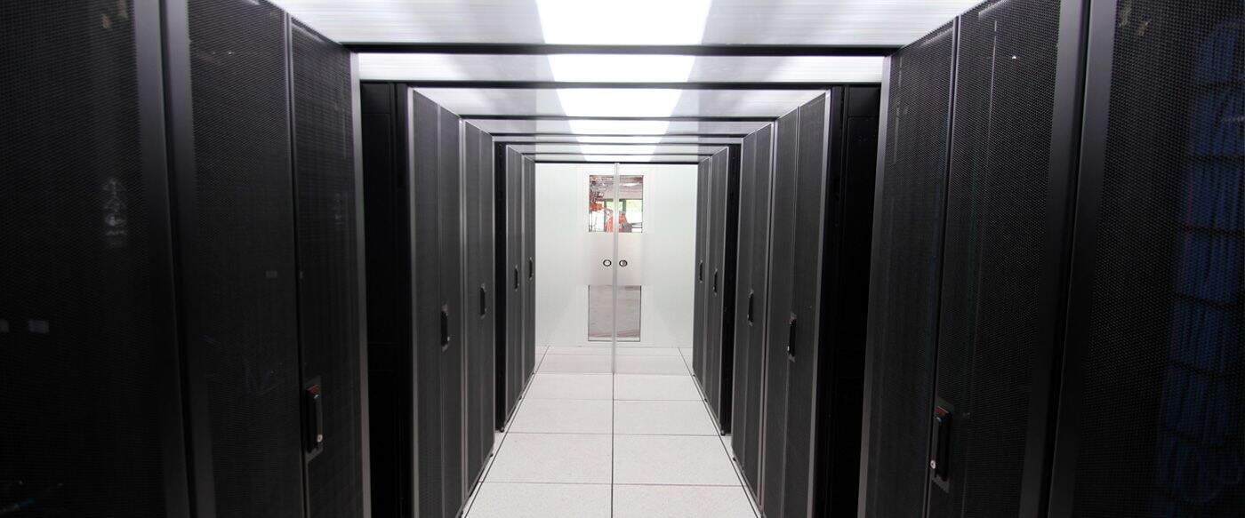 Allée d'armoires de serveurs noires sur un sol carrelé clair, éclairée par un plafond lumineux, créant une atmosphère technologique.