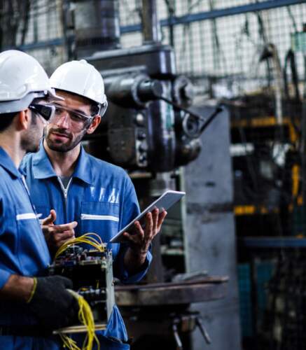 Deux techniciens industriels en tenue de travail et casque analysent un équipement électronique dans un atelier de production.