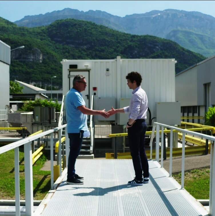 Deux techniciens se serrant la main devant un conteneur technique sur une plateforme industrielle extérieure.