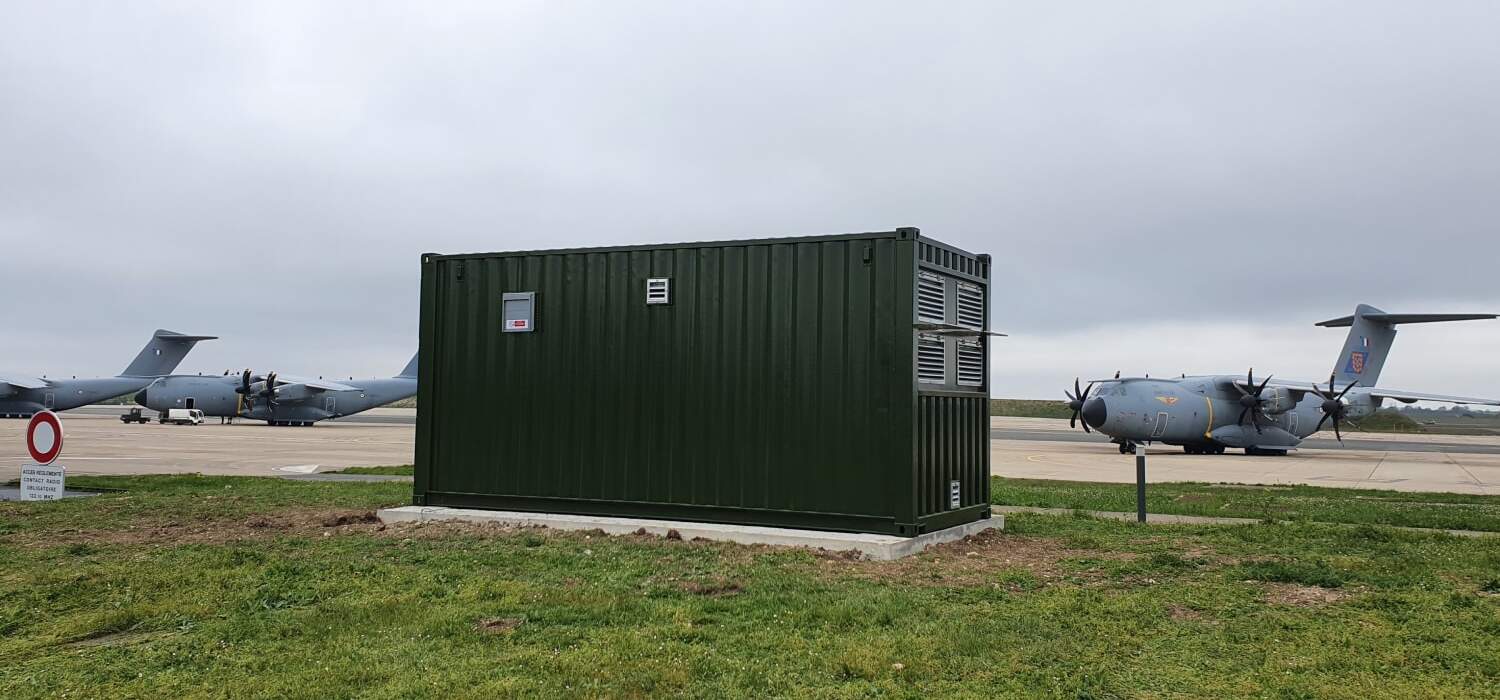 Conteneur technique installé sur une base aérienne, à proximité d’avions militaires sur le tarmac.