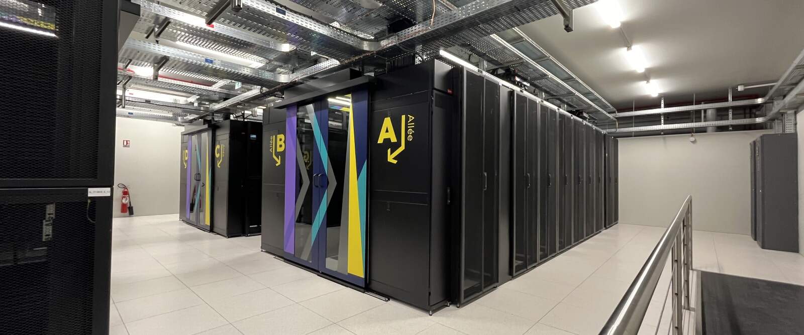 Salle informatique avec rangées de baies de serveurs noirs, allées identifiées A et B, câblage au plafond et sol technique dans un data center moderne.