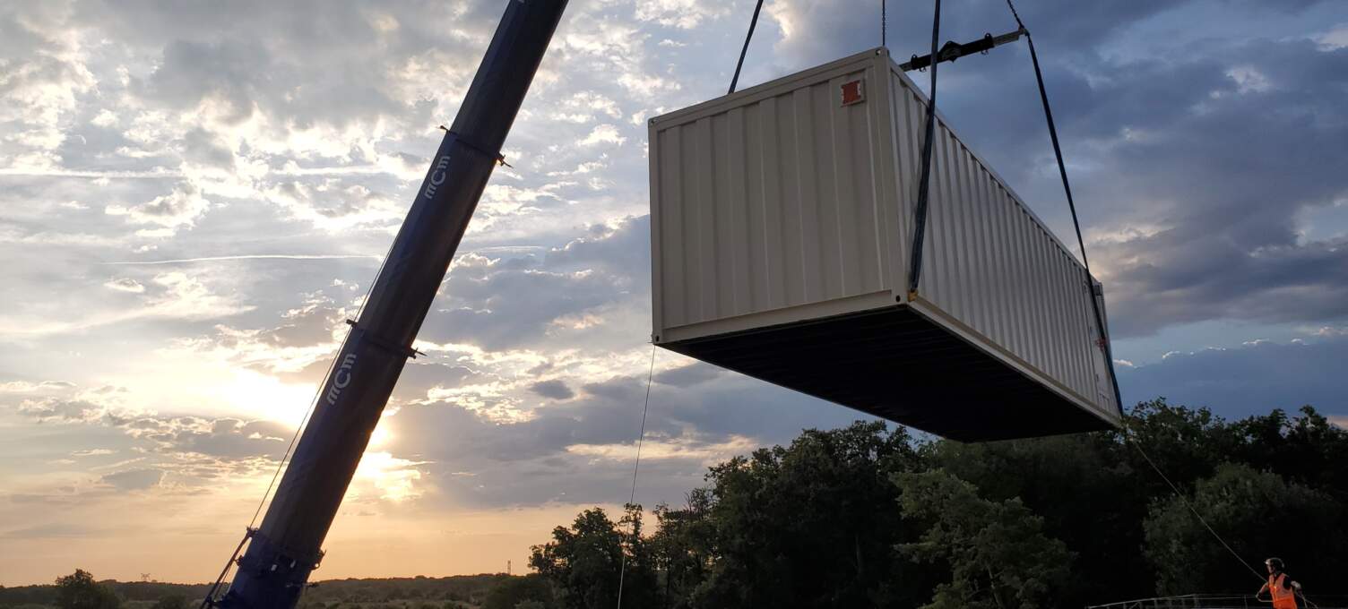 Un conteneur blanc est levé par une grue, avec un ciel nuageux et un coucher de soleil en arrière-plan. Des arbres verdoyants sont visibles.