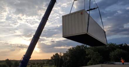 Un conteneur blanc est levé par une grue, avec un ciel nuageux et un coucher de soleil en arrière-plan. Des arbres verdoyants sont visibles.