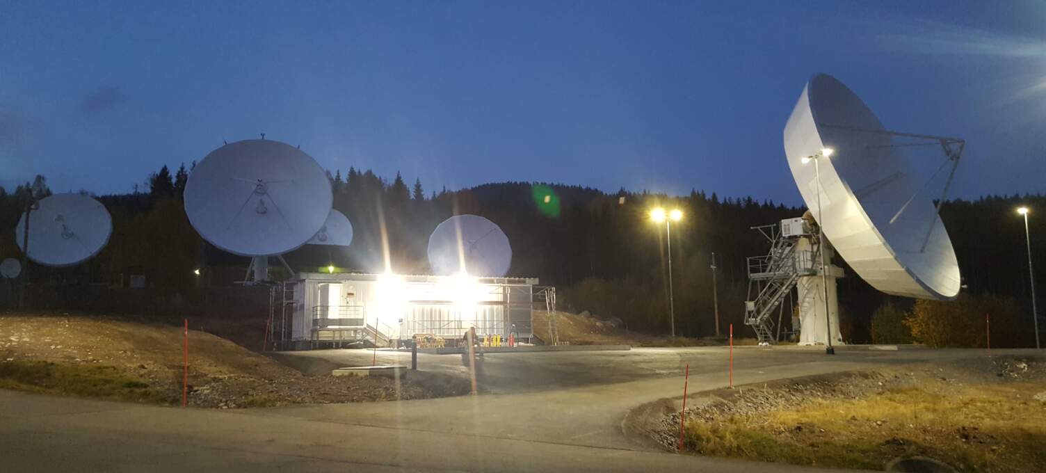 Parc de relais satellites au crépuscule, avec plusieurs antennes paraboliques blanches illuminées, entourées de forêt.