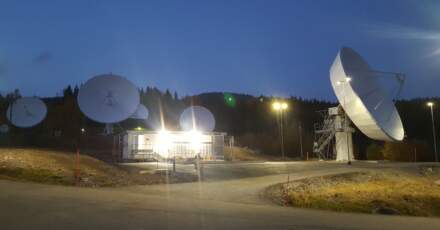 Parc de relais satellites au crépuscule, avec plusieurs antennes paraboliques blanches illuminées, entourées de forêt.