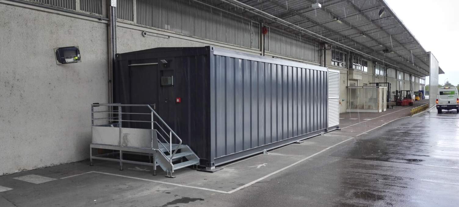 Conteneur métallique gris placé le long d'un bâtiment, avec des escaliers en métal et un sol en béton humide. Ambiance industrielle.