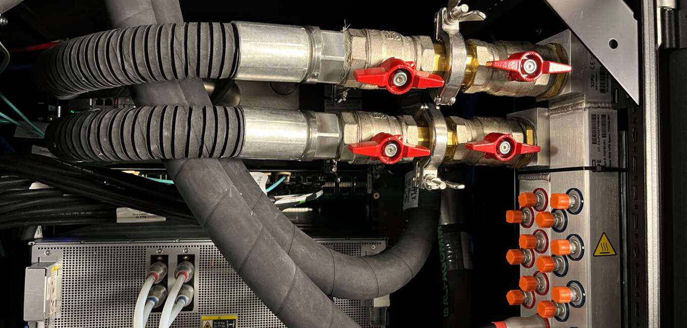 Panes de serveurs avec des tubes de refroidissement métalliques, valves rouges, câblage et boutons multicolores sur un fond noir.