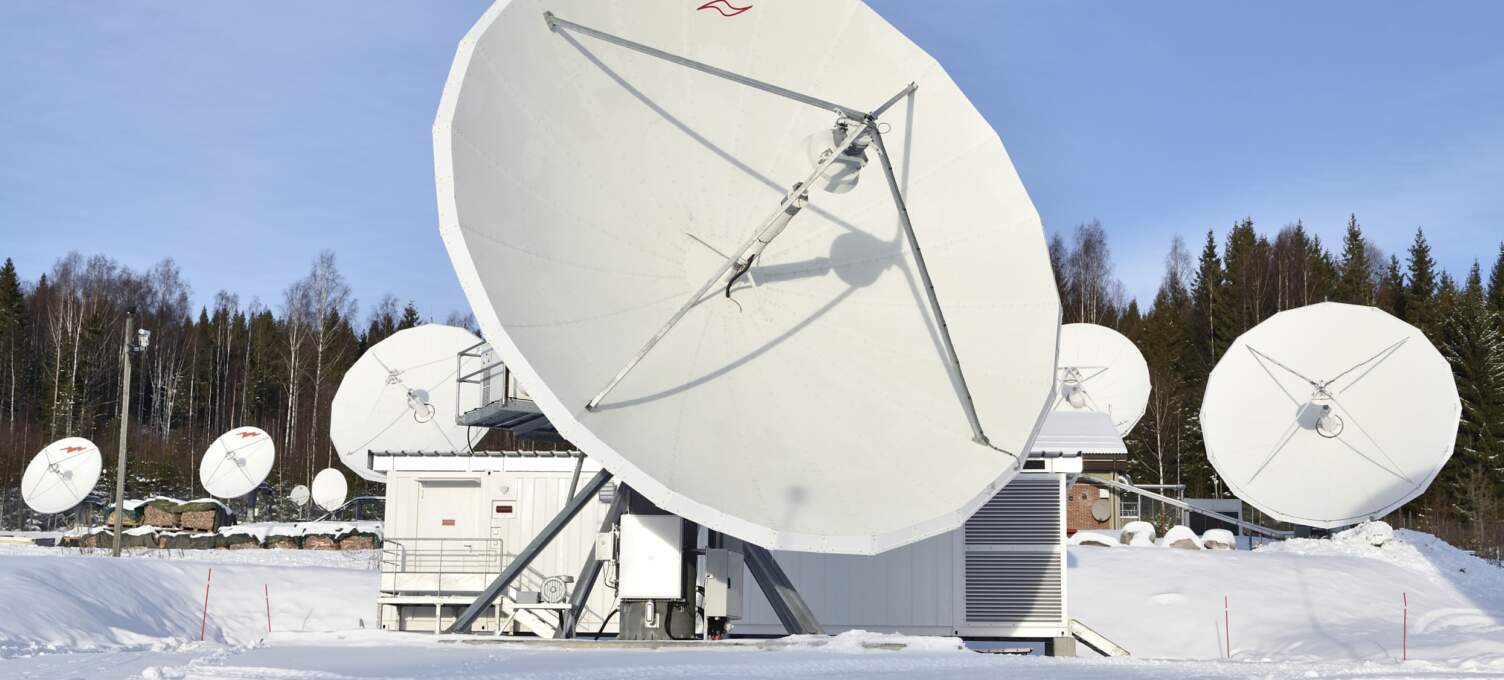 Antennes satellite blanches de grande taille sur un site enneigé, avec des arbres en arrière-plan et un bâtiment technique à proximité.