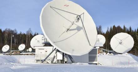 Antennes satellite blanches de grande taille sur un site enneigé, avec des arbres en arrière-plan et un bâtiment technique à proximité.