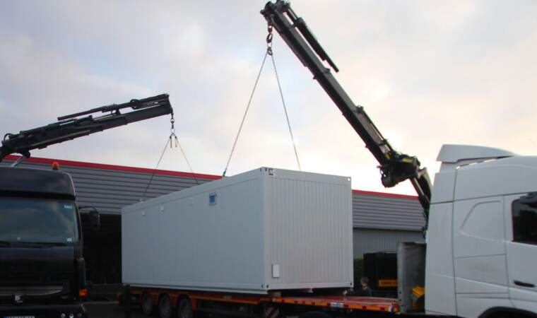 Camion équipé d’une grue soulevant un module préfabriqué blanc pour le déposer sur une remorque, sur un site industriel au lever ou coucher du jour.