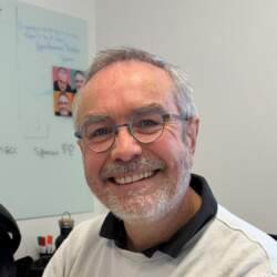 Homme souriant avec des lunettes et une barbe grise, assis à un bureau avec un tableau blanc en arrière-plan. Ambiance professionnelle.