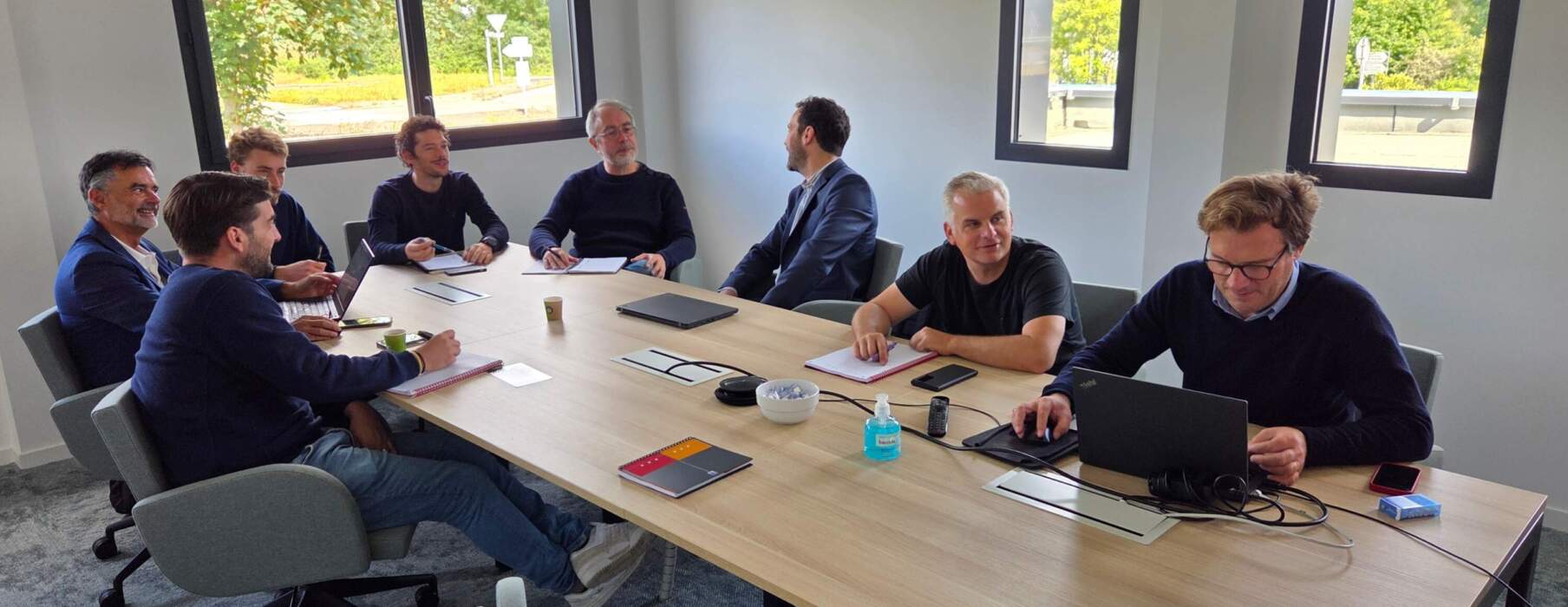 Réunion d’équipe autour d’une table de conférence dans un bureau lumineux, plusieurs collaborateurs échangent et travaillent sur des ordinateurs portables et des carnets.