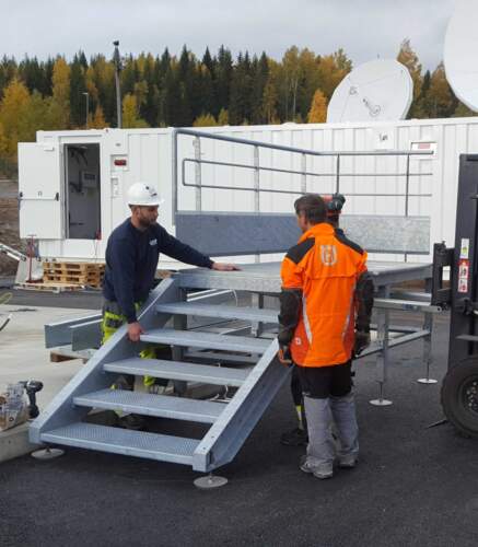 Techniciens installant une plateforme d’accès métallique devant un module technique sur un site industriel.