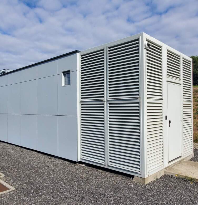 Bâtiment modulaire blanc avec façade à lattes, isolé sur un site herbeux, ciel nuageux en arrière-plan.