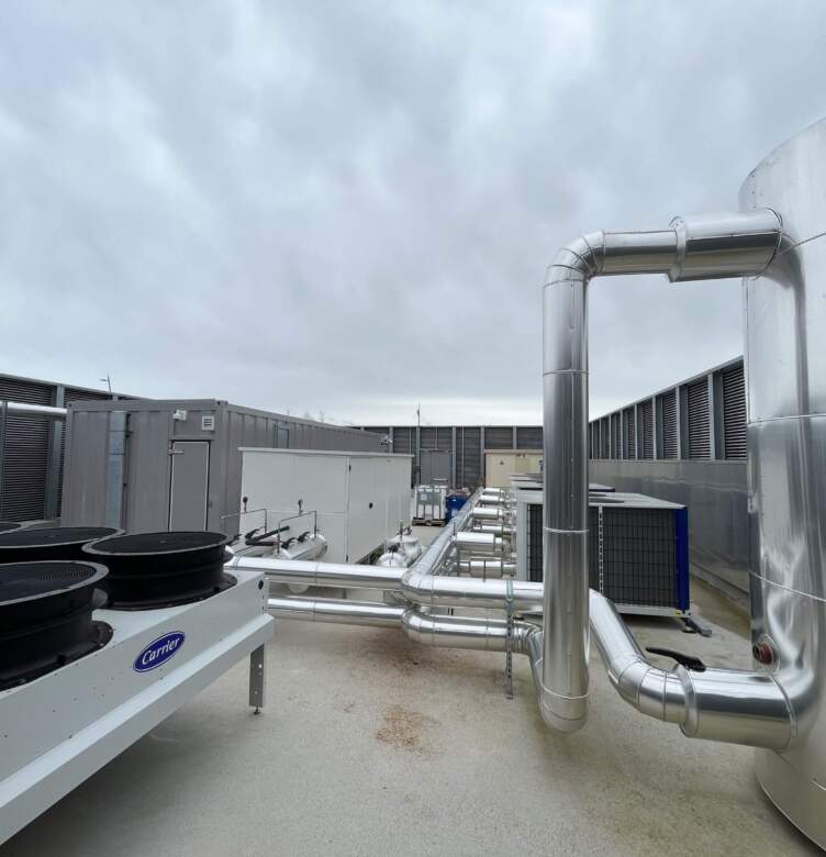 Système de refroidissement avec tuyauterie en métal et grandes unités sur le toit d'un datacenter, sous un ciel nuageux.