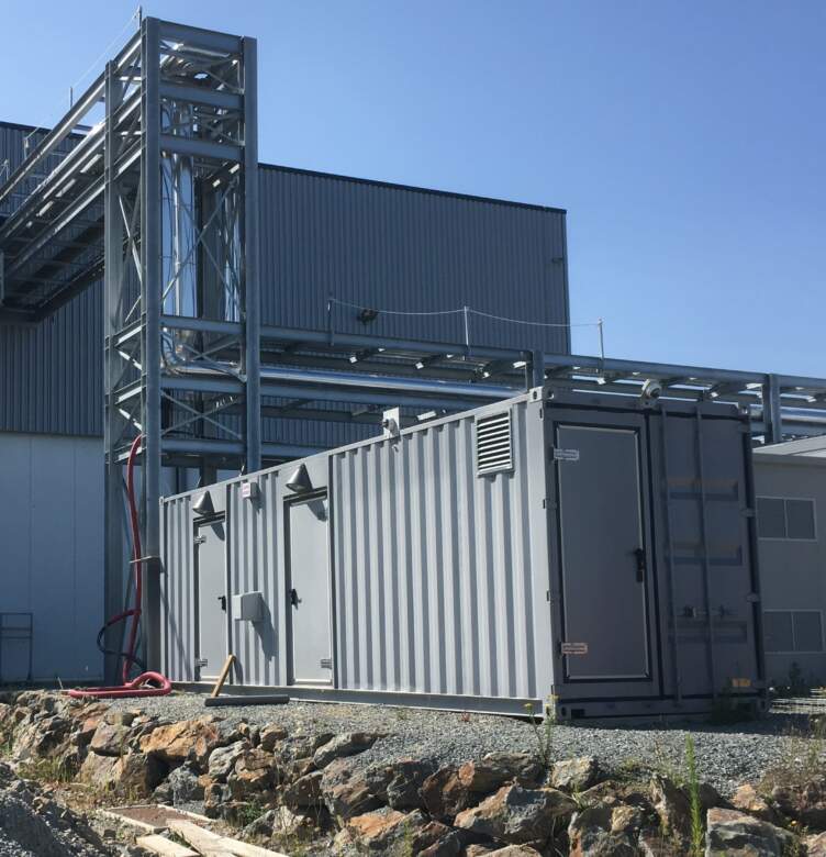 Conteneur gris modulaire installé près d'un bâtiment industriel, avec structures métalliques et tuyaux visibles sous un ciel bleu.