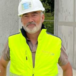 Homme souriant portant un gilet de sécurité fluorescent et un casque, devant une structure de bâtiment en béton.