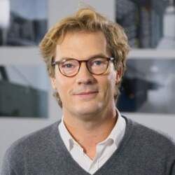 Homme souriant avec des lunettes et des cheveux bouclés, portant un pull gris et une chemise blanche, devant des images de datacenters.