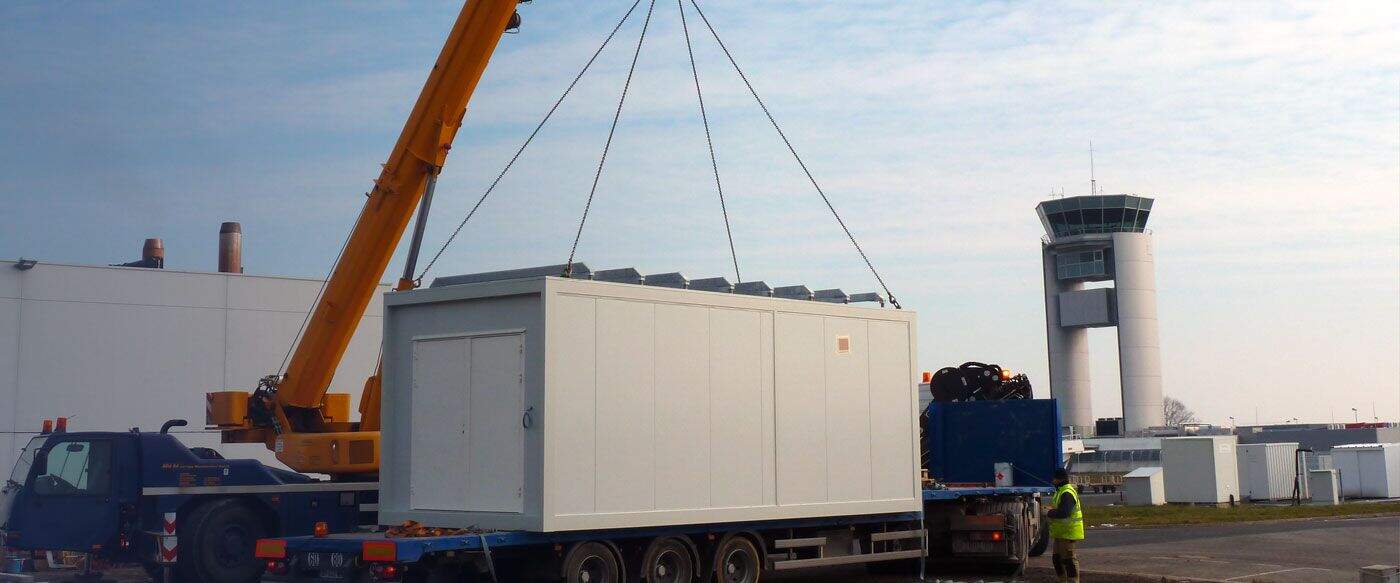 Grue jaune soulevant un conteneur blanc sur un camion à proximité d'une tour de contrôle d'aéroport sous un ciel clair.