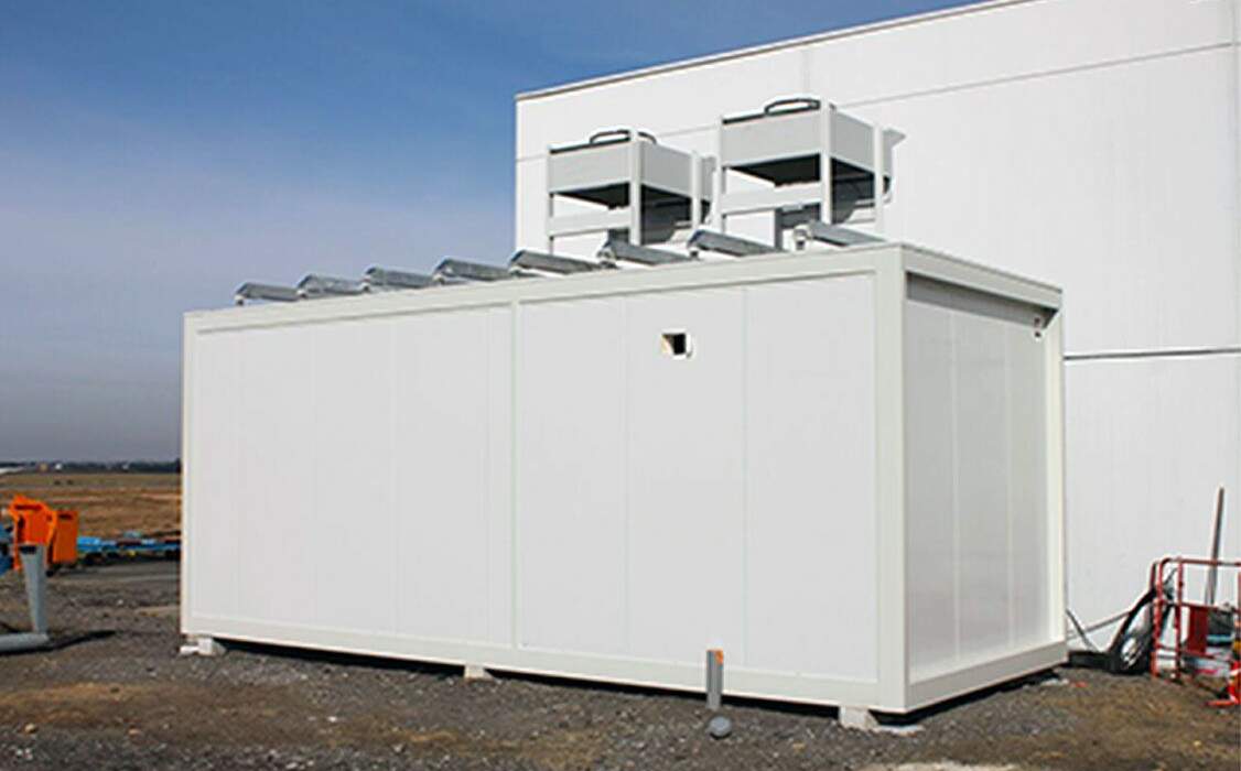 Cabine blanche de grande taille avec toits gris, située à l'extérieur, sur un sol terreux, sous un ciel clair.