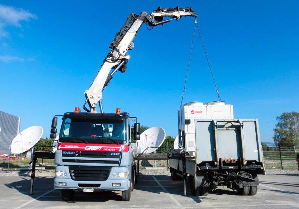 Camion-grue blanc avec bras télescopique soulevant un conteneur gris sur un terrain ensoleillé, entouré de satellites.