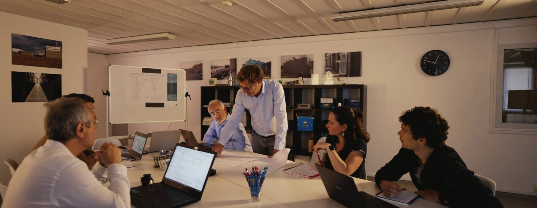 Réunion dans une salle avec six personnes autour d'une table, analysant des plans, ordinateurs portables ouverts, murs avec photos.