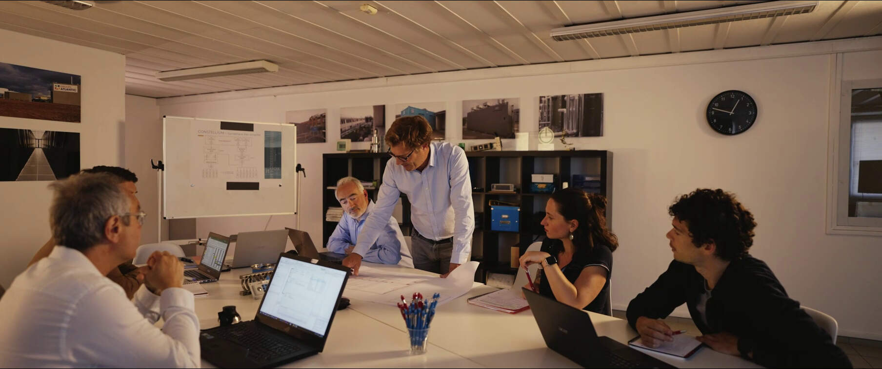 A meeting in a room with six people sitting around a table, reviewing plans, laptops open, and photos on the walls.