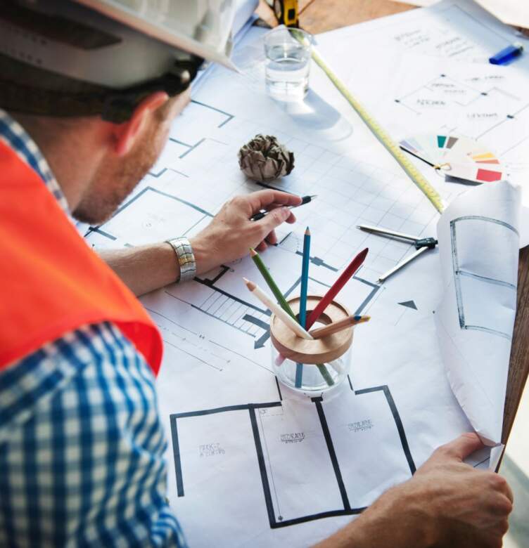 An engineer is examining blueprints on a table, surrounded by colored pencils, wearing a hard hat and an orange vest.