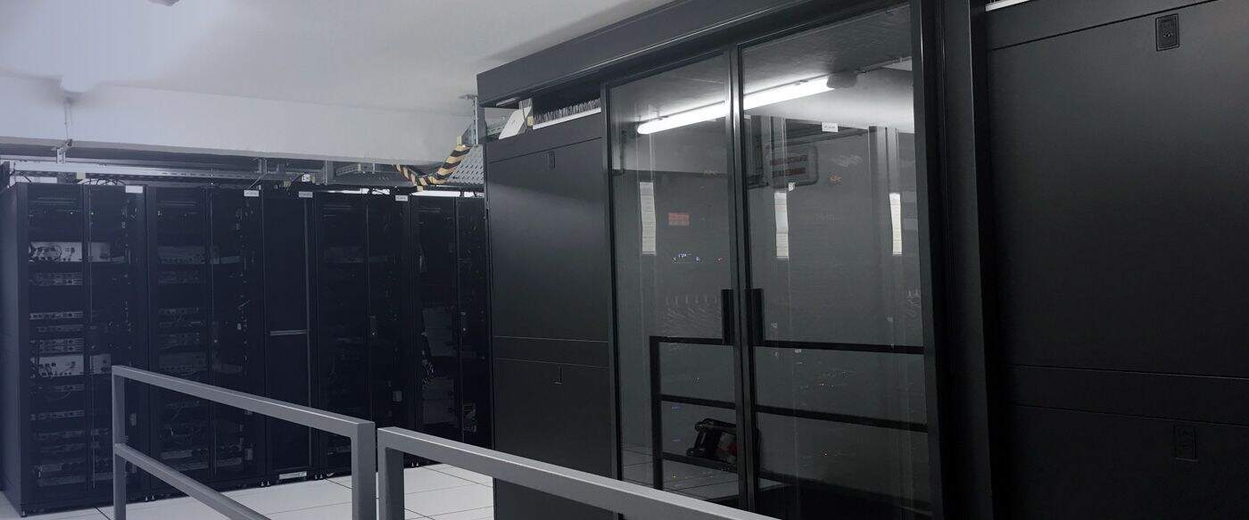 Centre de données avec plusieurs serveurs noirs alignés, une structure en verre au centre, éclairage fluorescent au plafond.