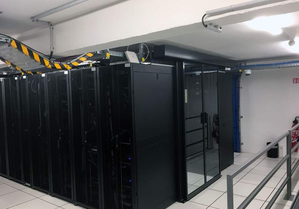 Salle de serveurs avec rangées de racks noirs, câblage en hauteur, sol blanc et éclairage fluorescent. Sécurisée et organisée.