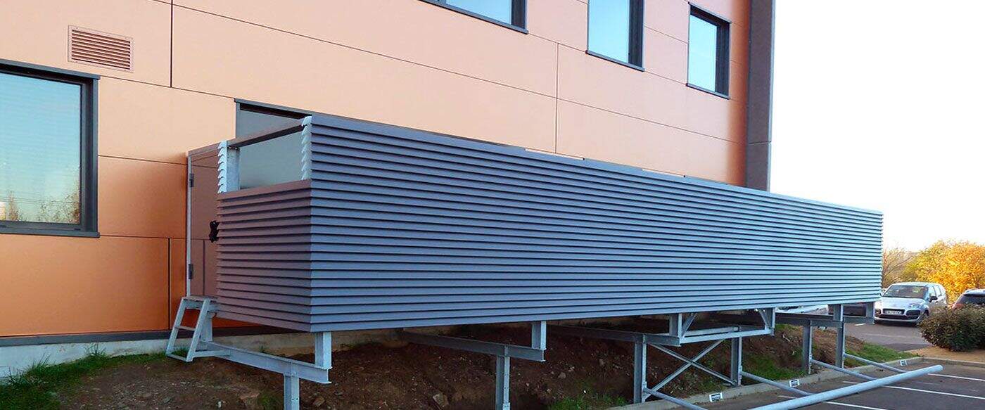 Cabine modulable en métal gris, posée sur un support surélevé, à côté d'un bâtiment aux murs orange. Ambiance extérieure claire.