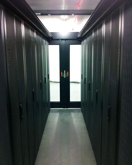 Allée étroite entre des rangées de serveurs noirs, menant à une porte double à poignées rouges sur fond blanc lumineux.