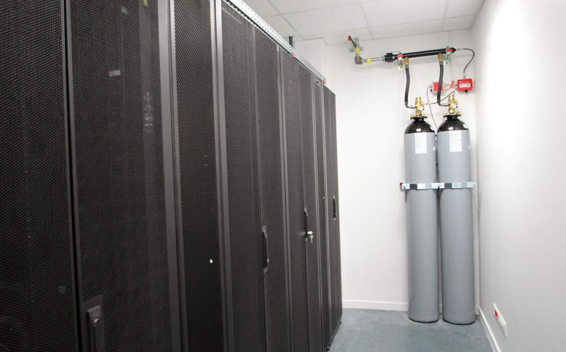 Salle technique équipée de plusieurs serveurs rackés noirs, deux grandes bouteilles grises et des tuyaux visibles sur le mur.